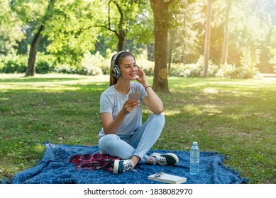 Young Happy Woman Sitting In The Park Surfing The Song Play List In Her Phone And Listening The Music On The Wireless Headphones