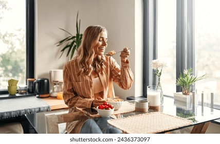 Young happy woman sitting at the table in the kitchen, eating cereal with milk, strawberries and drinking coffee. A woman is having breakfast in a cozy kitchen. - Powered by Shutterstock