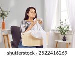 Young happy woman smelling fresh laundry aroma basket at home, clean clothes, white linen, housewife or maid spring cleaning with detergent, cleaner scent on fresh fabric, comfort sniff after washing 