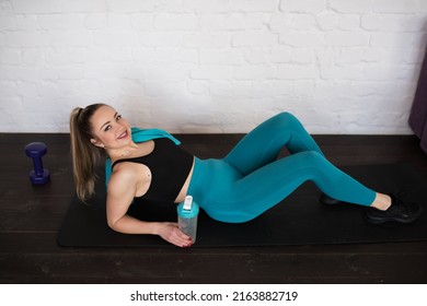 Young Happy Woman In Sportswear And A Towel Around Her Neck, Holding A Bottle Of Water In Her Hand On A Black Exercise Mat, Home Workout, Healthy Lifestyle. High Quality Photo