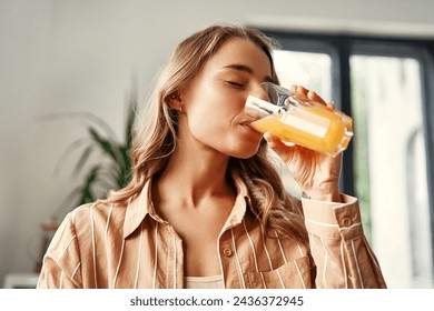 A young happy woman standing in the kitchen drinks orange juice from a glass. Woman having breakfast in a cozy kitchen. - Powered by Shutterstock
