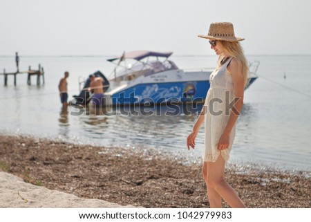 Young happy woman in a summer dress, sunglasses with a straw hat walking on a sea background, where you can see a pier and a boat. Vacation. Travel. 