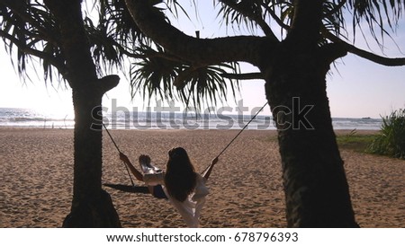 Young happy woman in swimsuit and shirt relaxing at swing at tropical ocean beach. Beautiful girl sitting on swing and enjoying summer vacation or holiday. Female having fun at shore. Rear back view.