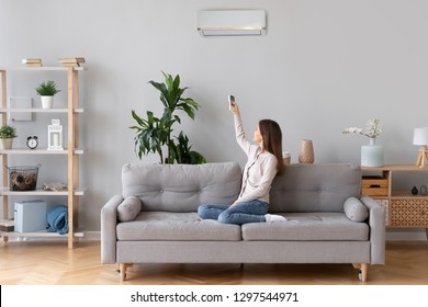 Young happy woman switching on air conditioner sitting on couch at convenient cozy home, lady relaxing on sofa in living room holding remote climate control to cooler system set comfort temperature - Shutterstock ID 1297544971