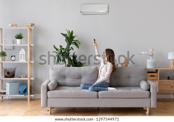 快適な家庭のソファに座るエアコンをつけ 居間のソファでくつろぐレディー リビングルームのリモコンを持つリビングルームのレディー より涼しいシステムで快適な温度を設定する の写真素材 今すぐ編集