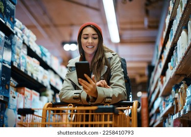 Young happy woman texting on cell phone while buying in supermarket. - Powered by Shutterstock