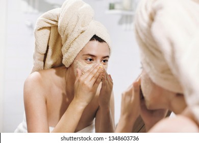 Young happy woman in towel making facial massage with  organic face scrub and looking at mirror in stylish bathroom. Girl applying scrub cream, peeling and cleaning skin. Skin Care