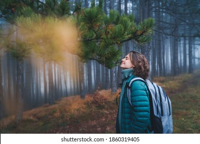 Young happy woman traveler in green jacket with backpack in foggy pine forest enjoying walking breathing fresh air