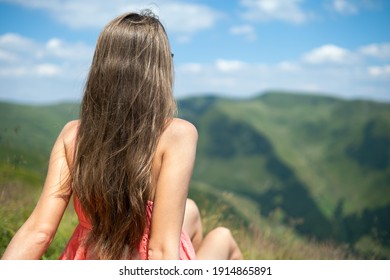 Young happy woman traveler in red dress resting on green grassy hillside on a windy day in summer mountains enjoying view of nature. - Shutterstock ID 1914865891