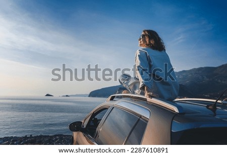 Young happy woman traveler sitting on car roof enjoying sea and sunset view. Summer vacation and adventure concept