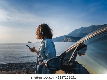 Young happy woman traveling on the sea, standing next to the car with a smartphone in her hands on the beach