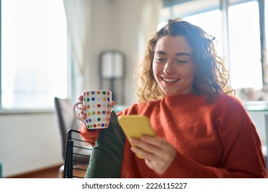 Young happy woman using cell phone drinking coffee relaxing at home. Smiling pretty lady holding smartphone enjoying mobile shopping, checking social media messages, playing game or buying online. - Powered by Shutterstock