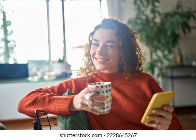 Young happy woman using cell phone drinking coffee relaxing at home. Smiling pretty lady holding smartphone enjoying mobile shopping, daydreaming checking news on smartphone in cozy morning. - Powered by Shutterstock