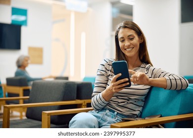 Young happy woman using cell phone in waiting room at doctor's office. Copy space. - Powered by Shutterstock