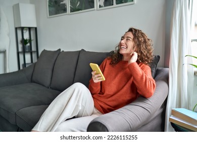 Young happy woman using mobile cell phone sitting on couch at home. Smiling cheerful lady laughing holding smartphone having fun while buying in ecommerce shop or watching funny videos online. - Powered by Shutterstock