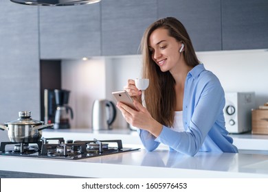 Young Happy Woman Using Smartphone And Wireless Headphones For Reading Audio Book During Coffee Break In The Kitchen At Home. Modern Mobile People 