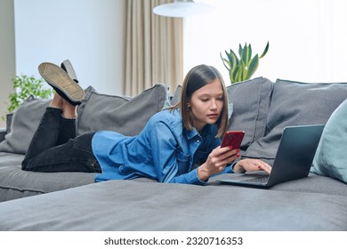 Young happy woman using smartphone laptop, lying on sofa at home - Shutterstock ID 2320716353