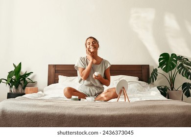 A young happy woman, waking up in the morning, sits on her bed, applying moisturizer to her face. Skin care for face and body, morning routine, spa treatments. - Powered by Shutterstock