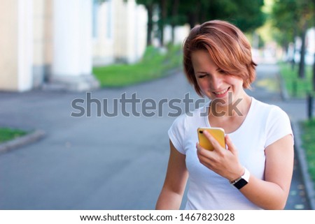 A young happy woman is walking around the city and looking at her phone and smiling. Girl in a white T-shirt and modern technology: smartphone and smartwatch. Making a video call