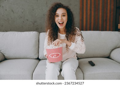 Young happy woman wear casual clothes sits on grey sofa couch eat popcorn watch movie film look camera stay at home hotel flat rest relax spend free spare time in living room indoor. Lounge concept - Shutterstock ID 2367234527