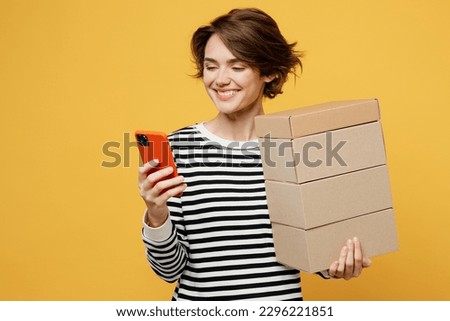 Young happy woman wear casual striped black and white shirt hold in hand stack cardboard blank boxes use mobile cell phone isolated on plain yellow color background studio portrait. Lifestyle concept