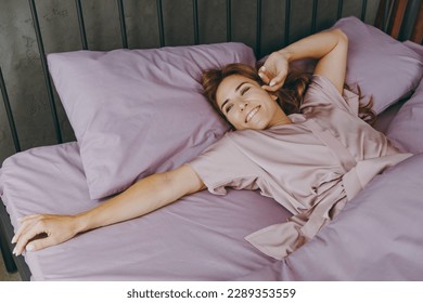 Young happy woman wear purple t-shirt pajama lying in bed look aside rest relax spend time in bedroom lounge home in own room hotel wake up dream be lost in reverie good mood day. Real estate concept