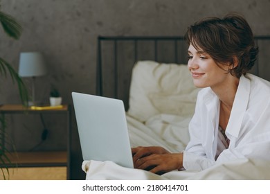 Young happy woman wear white shirt pajama she lying in bed hold use work on laptop pc computer study online browsing internet rest relax spend time in bedroom lounge home in own room hotel wake up.