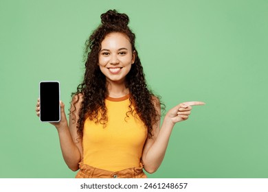 Young happy woman wear yellow tank shirt top hold in hand use mobile cell phone with blank screen workspace area point index finger aside isolated on plain pastel green background. Lifestyle concept - Powered by Shutterstock