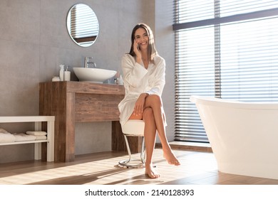 Young Happy Woman Wearing Silk Robe Talking On Mobile Phone In Modern Bathroom, Beautiful Smiling Lady Sitting On Chair And Having Pleasant Conversation On Cellphone At Home, Copy Space