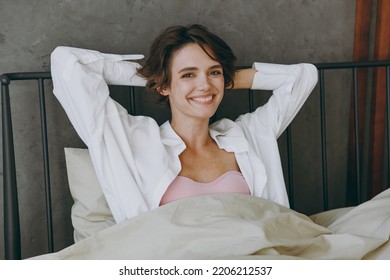 Young happy woman wearing white shirt pajama she lying in bed hold hands behind neck rest relax spend time in cosy bedroom lounge home in own room hotel wake up dream be lost in reverie good mood day. - Shutterstock ID 2206212537