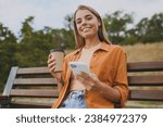 Young happy woman wears orange shirt casual clothes use mobile cell phone drink coffee look camera sit on bench walk rest relax in spring green city park outdoors on nature. Urban lifestyle concept