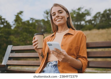 Young happy woman wears orange shirt casual clothes use mobile cell phone drink coffee look camera sit on bench walk rest relax in spring green city park outdoors on nature. Urban lifestyle concept - Powered by Shutterstock
