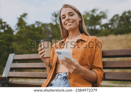 Young happy woman wears orange shirt casual clothes use mobile cell phone drink coffee look camera sit on bench walk rest relax in spring green city park outdoors on nature. Urban lifestyle concept