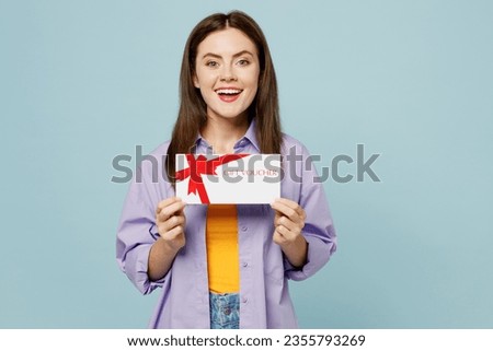 Young happy woman wears purple shirt yellow t-shirt casual clothes hold gift certificate coupon voucher card for store isolated on plain pastel light blue background studio portrait. Lifestyle concept
