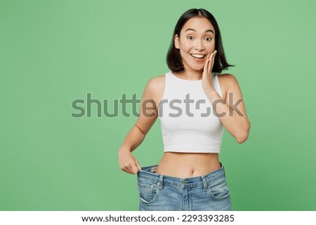Young happy woman wears white clothes show loose pants on waist after weightloss hold face isolated on plain pastel light green background. Proper nutrition healthy fast food unhealthy choice concept
