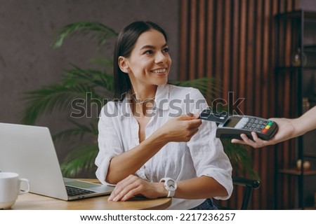 Young happy woman in white shirt hold pay waiter hold wireless bank payment terminal process acquire credit card sit at table in coffee shop cafe restaurant indoor work or study on laptop pc computer