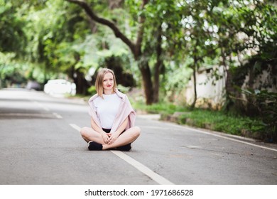 Young happy woman in white t-short is sitting in the middle of the asphalt road with greens on background