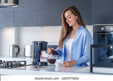 Young Happy Woman With White Wireless Earphones Listening Music And Audio Book During Making Fresh Aromatic Coffee Using Coffee Maker In The Kitchen At Home. Modern Mobile People 