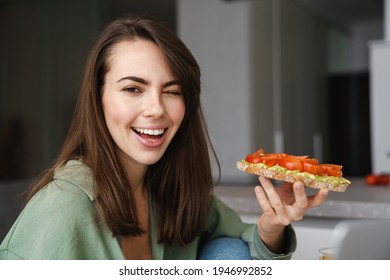 Young Happy Woman Winking While Eating Avocado Toast At Home Kitchen