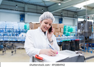 Young Happy Woman Worker In Factory Checking Robotic Line For Water Bottling And Packing. Inspection Quality Control And Talking On Her Cell Phone. People At Work.