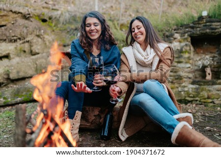 young happy women drinking glass of red wine. Females warming next to the fire. Campfire, outdoors activities concept.