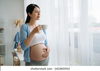 young healthy asian pregnant woman in sports bra and leggings standing by window touching belly as if hugging future baby. mom enjoy drink black coffee at home to relax and stay fit look view window