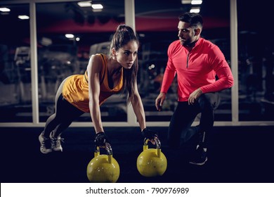 Young healthy focused sporty strong shape girl with a ponytail doing push ups exercises with the kettlebells while handsome helpful personal trainer crouching next to her in the gym. - Powered by Shutterstock