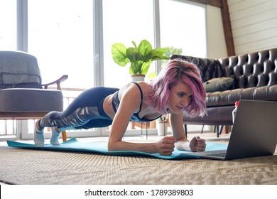 Young healthy sporty fit woman with pink hair wear sportswear doing plank sport training exercise watching online class tutorial on laptop at home. Online fitness workout video virtual coach concept.