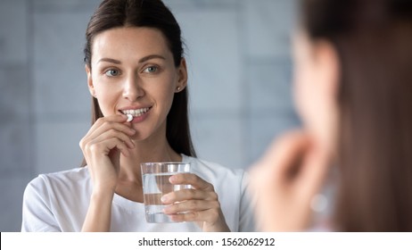 Young healthy woman holding pill glass of water looking in mirror, adult lady take daily medicine diet vitamin omega supplements for beauty skin hair health care medicament stand in bathroom concept - Shutterstock ID 1562062912
