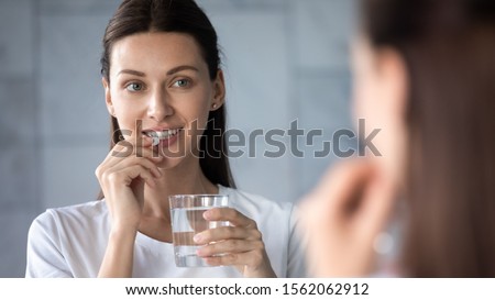 Young healthy woman holding pill glass of water looking in mirror, adult lady take daily medicine diet vitamin omega supplements for beauty skin hair health care medicament stand in bathroom concept