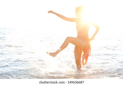 Young Healthy Woman In The Sea Shore, Kicking The Ocean Water With Her Legs While Dressed And With The Sun Rays And Flare Covering Her Face And Features. Golden Silhouette.