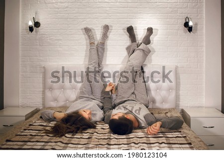 A young heterosexual couple is lying on the bed with their legs up against the wall and holding each other's hands. Lovers relax at home. Beautiful young couple spend time together