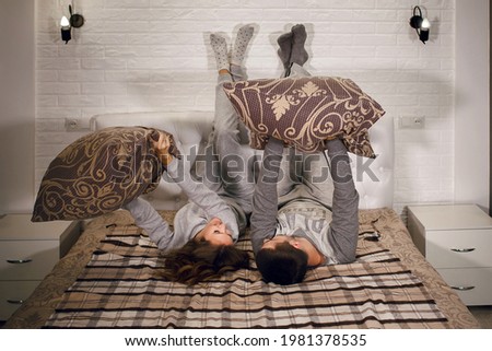 A young heterosexual couple is lying on the bed with their feet against the wall and pillow fights. Lovers are resting at home. Beautiful young couple have fun and enjoy communicating with each other