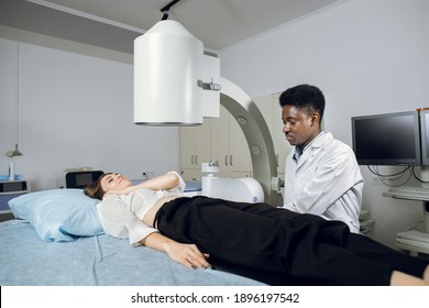 Young high-skilled focused black man doctor in white coat, providing lithotripsy procedure for his lying female patient with modern ultrasonic lithotriptor to break up stones